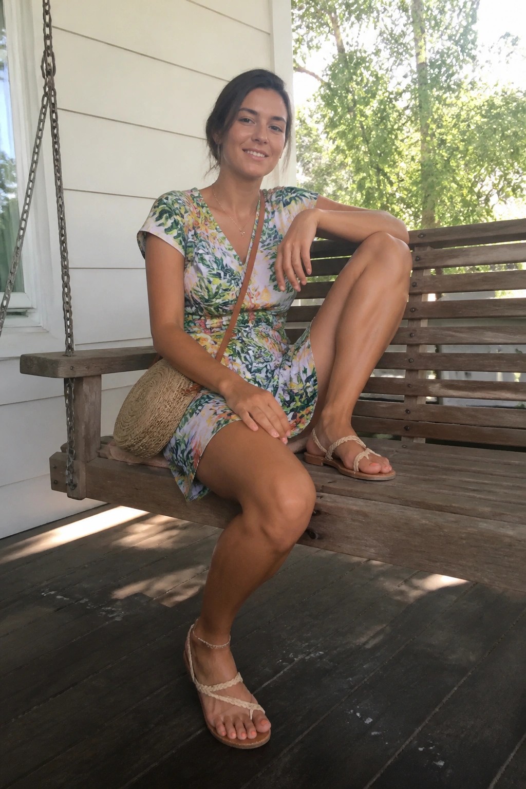 Smiling woman in floral dress seated on wooden porch swing with bag