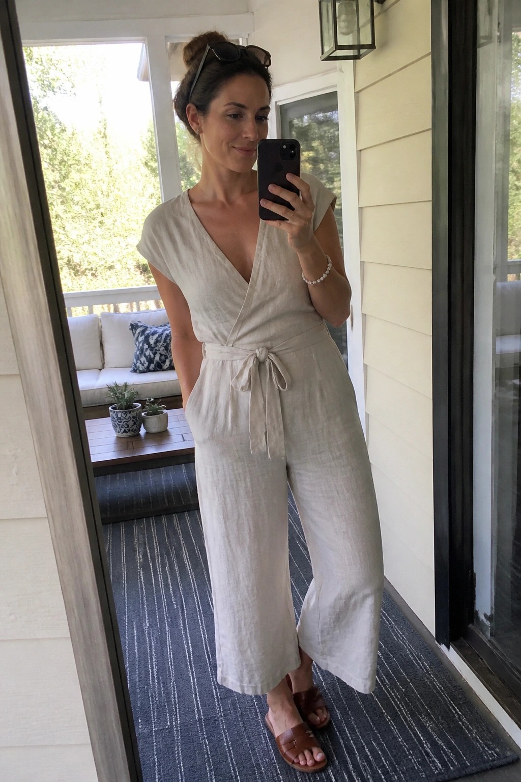 Woman in beige linen jumpsuit taking mirror selfie on porch.