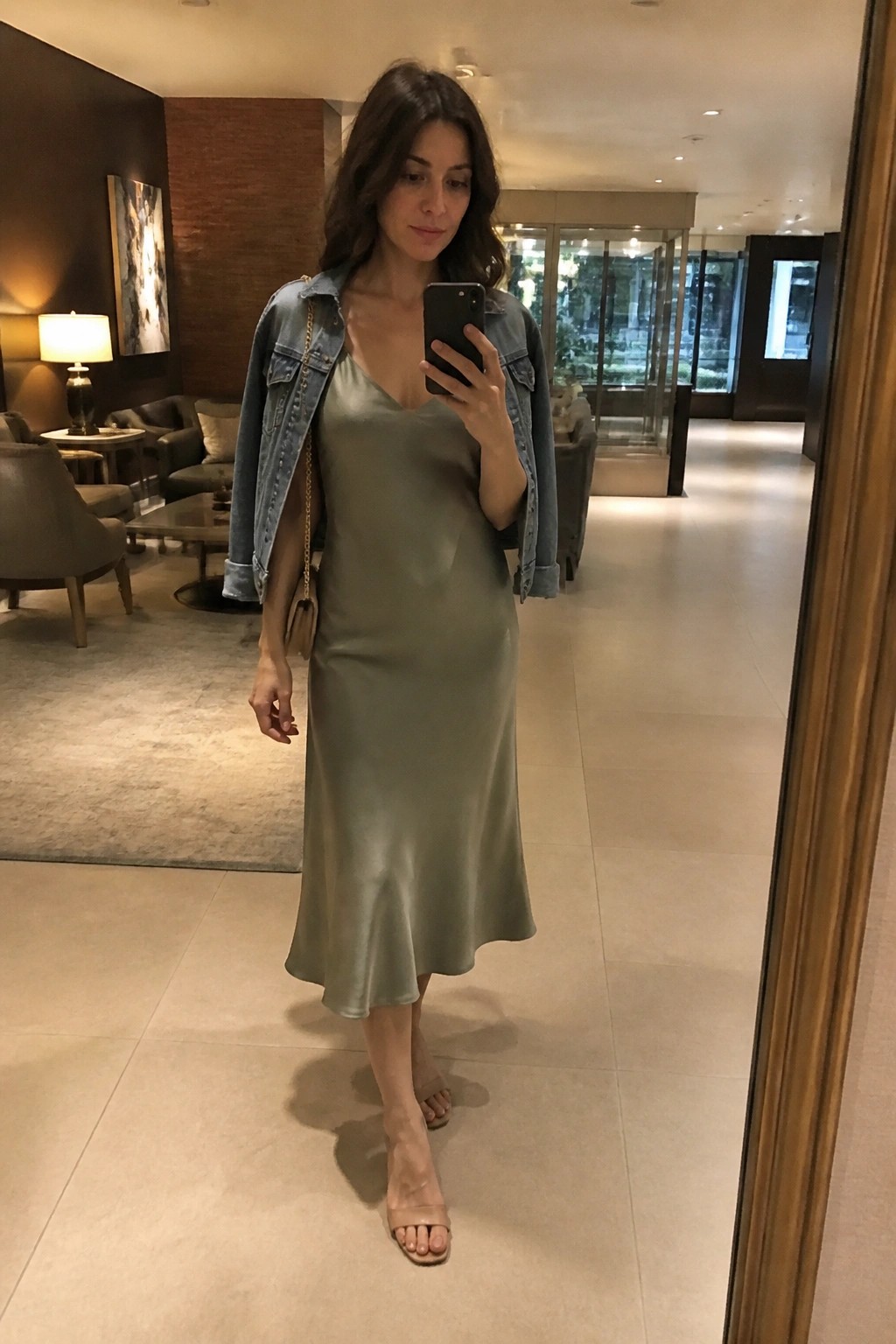 Woman in sage green satin slip dress with blue denim jacket layered over shoulders, chain strap bag, and nude heels holding a phone.