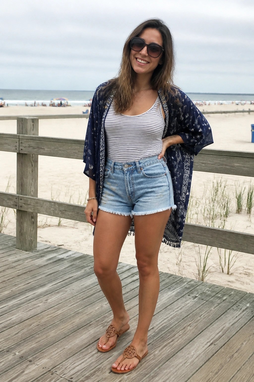Woman smiling on beach boardwalk in striped top, denim shorts, blue kimono