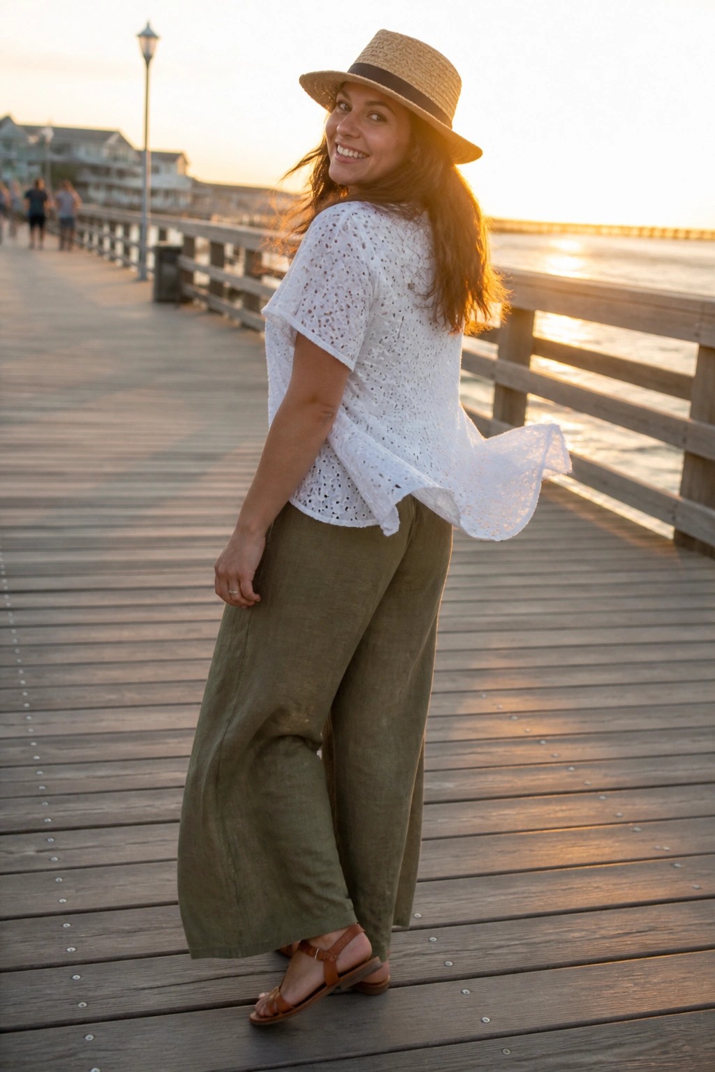 Woman in white eyelet short sleeve top, olive wide leg pants, straw hat and brown sandals on wooden pier.