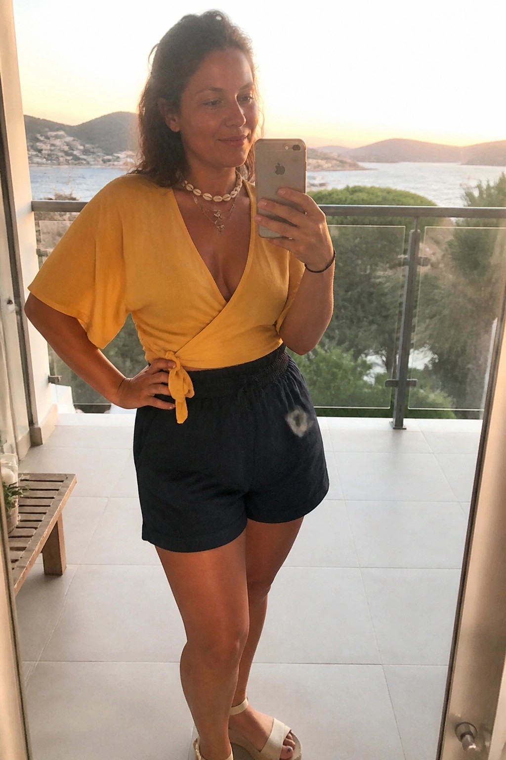 Woman in yellow wrap top and navy shorts takes balcony selfie overlooking sunset sea.