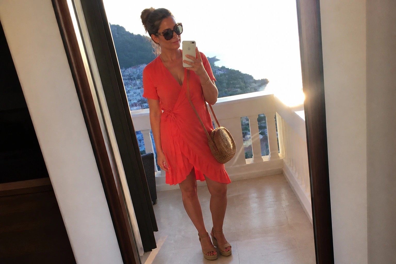 Woman in a bright coral short sleeve wrap mini dress with waist tie ruffled hem platform wedge sandals and round straw bag posing for a mirror selfie.