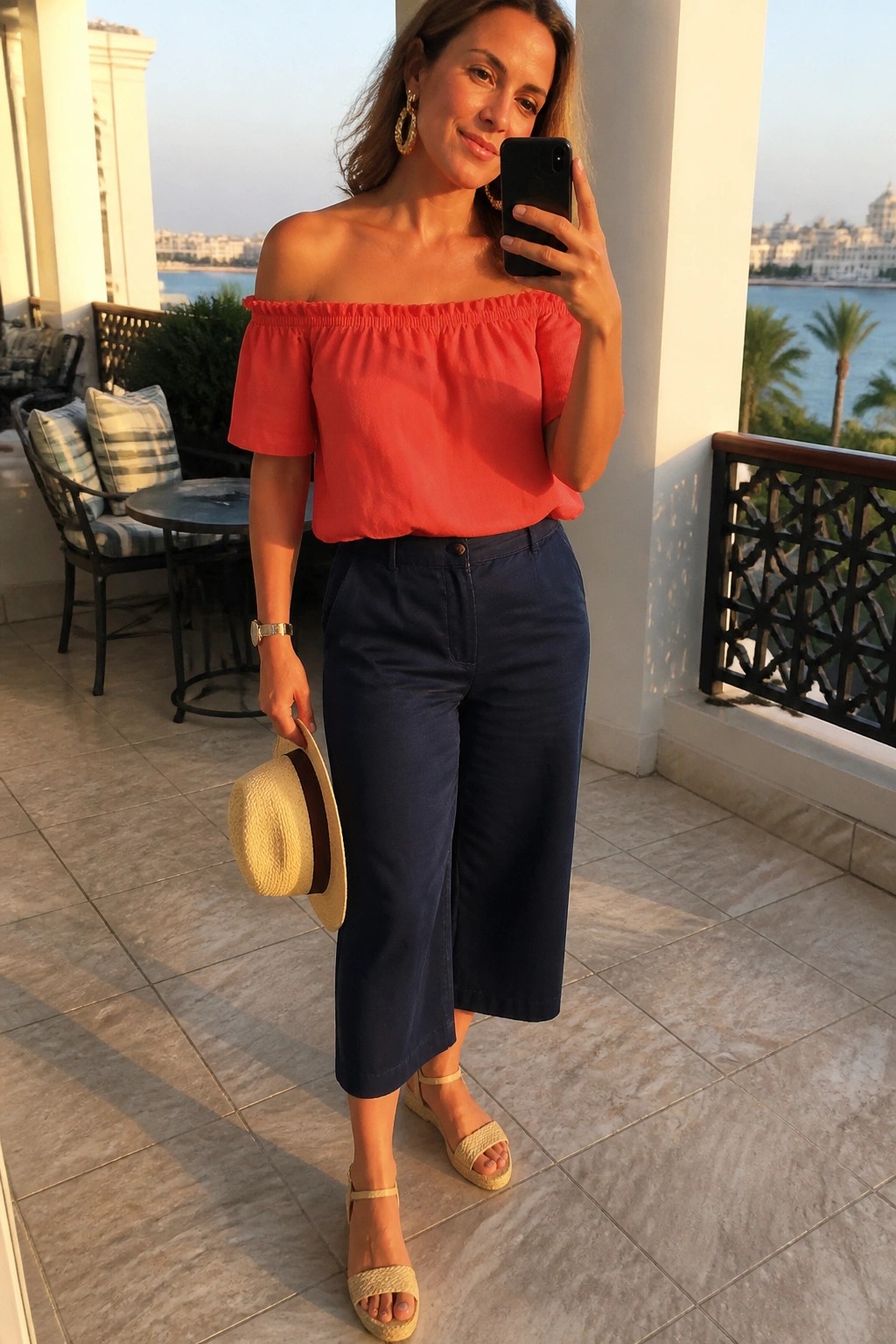 Woman wearing red off shoulder blouse with navy wide leg cropped pants beige wedge sandals and holding a straw hat while taking a mirror selfie.