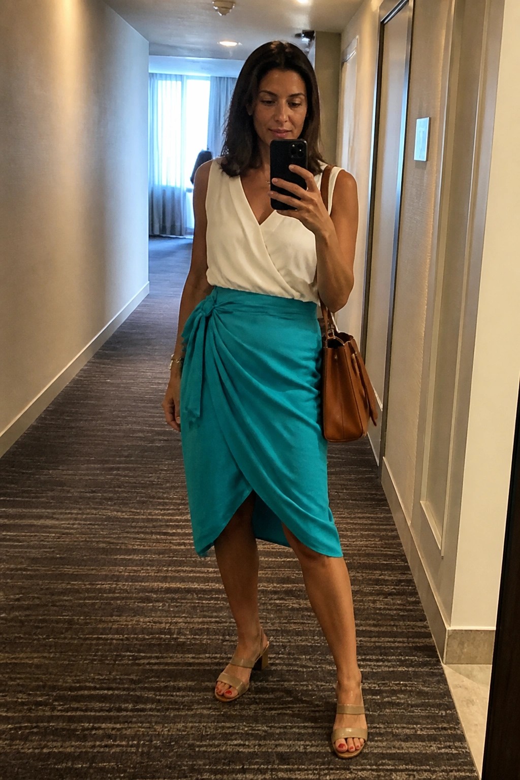 Woman in white top and teal wrap skirt taking mirror selfie in hallway