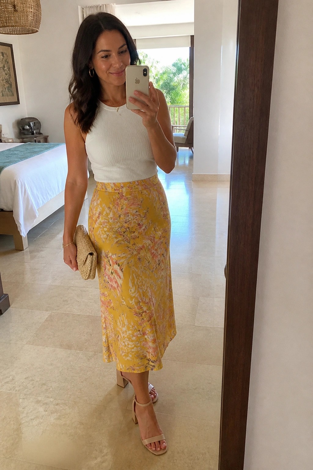 Woman in white top and yellow floral skirt taking mirror selfie in bedroom