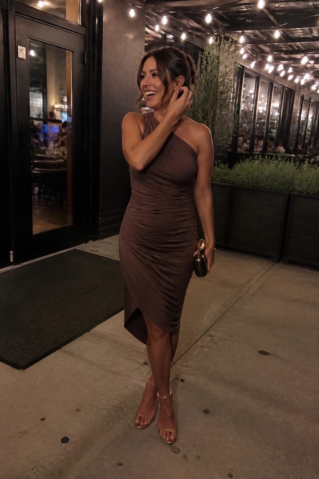 Woman in a brown one-shoulder ruched midi dress with asymmetrical hem paired with gold strappy heels and a small gold clutch.