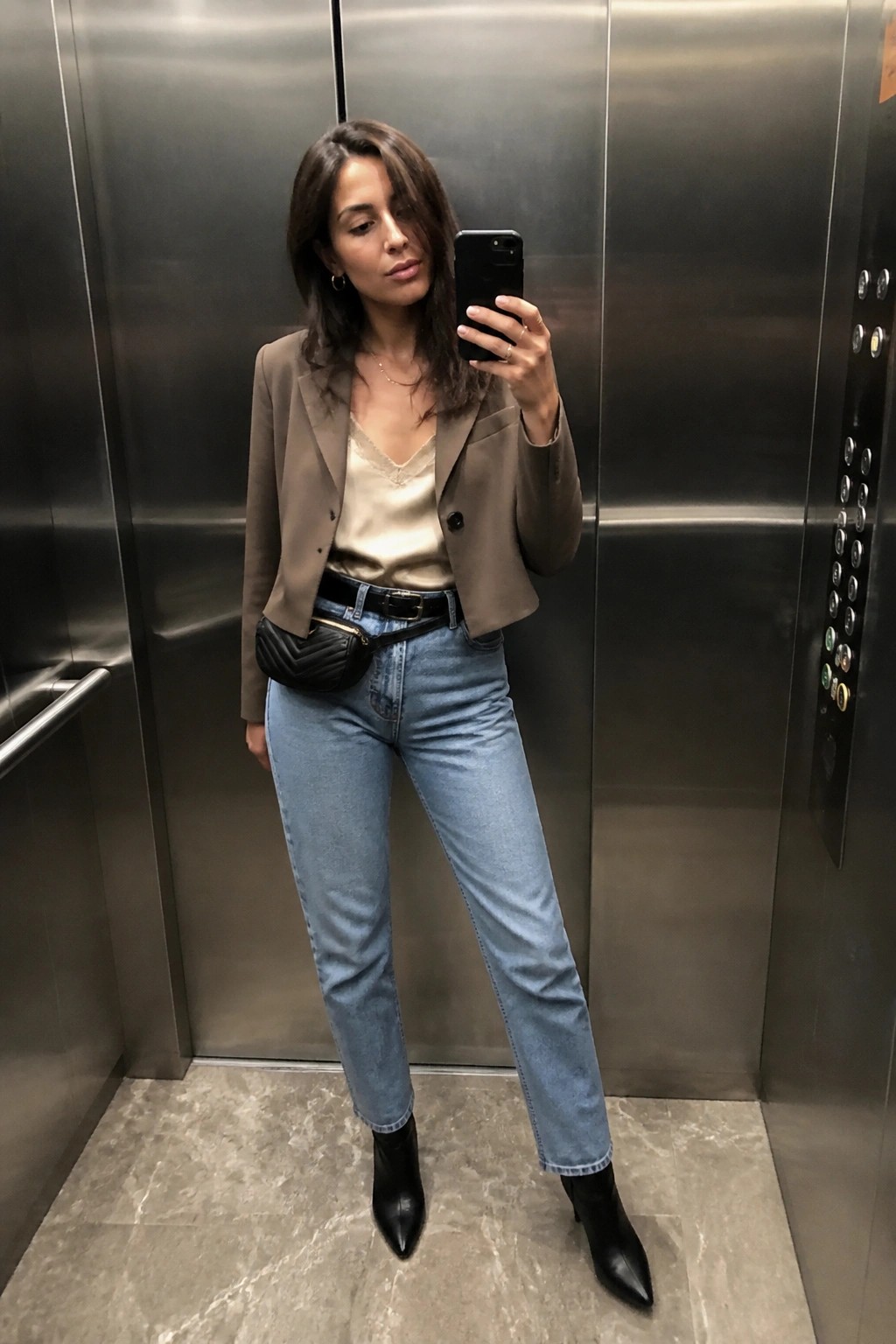 Woman in taupe blazer and jeans taking mirror selfie in elevator.