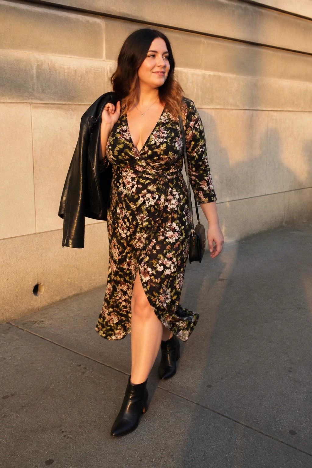 A midsize woman in a black floral wrap midi dress with deep V neckline, three quarter sleeves, front slit, layered with black leather jacket over shoulder, black ankle boots, and small black crossbody bag.
