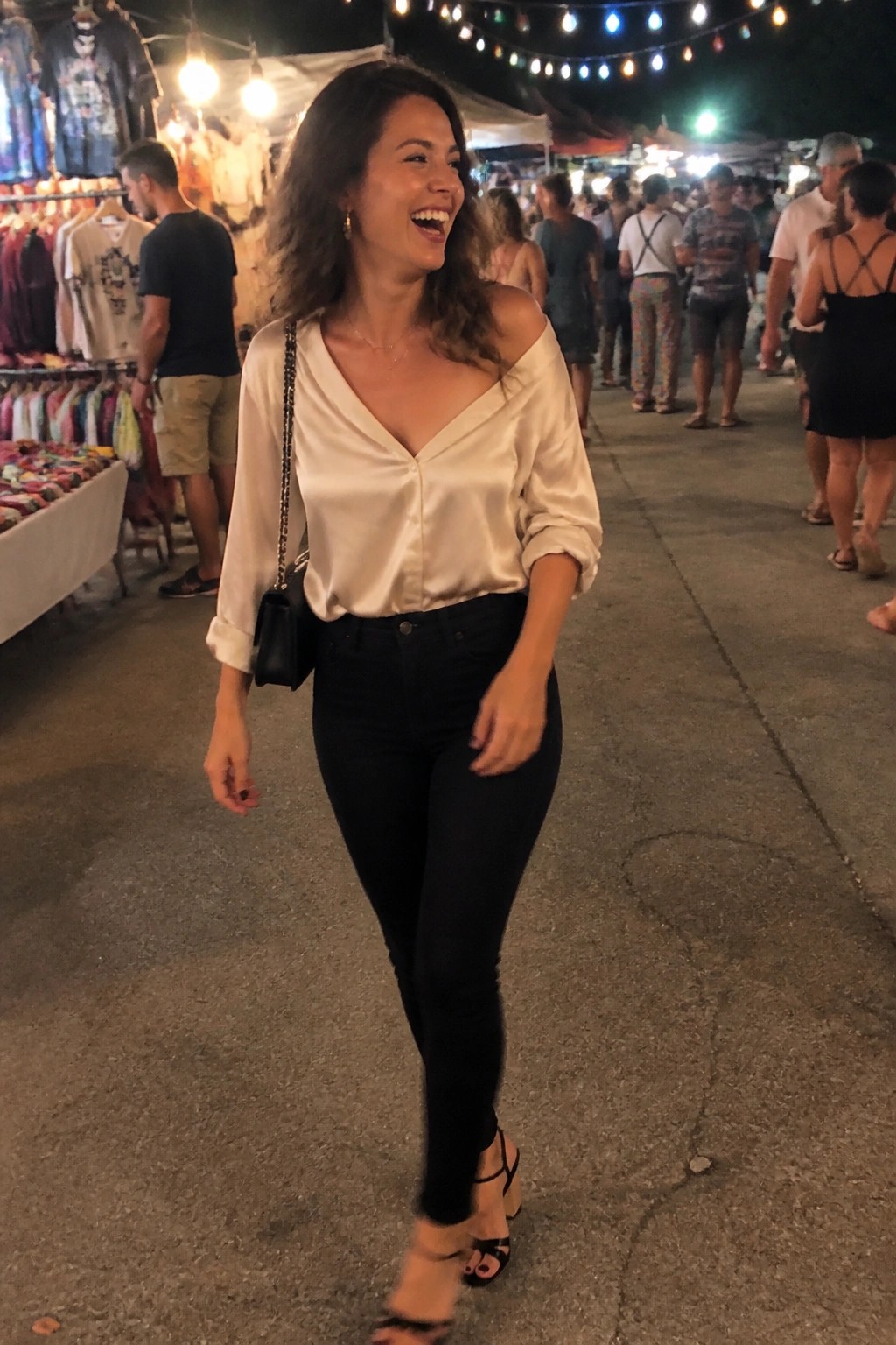 Woman laughing in satin blouse and jeans at crowded night market with lights