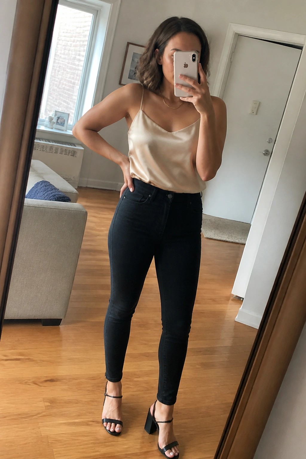 Woman wearing a beige satin camisole tucked into black skinny jeans with black block heel sandals and a thin gold necklace while taking a mirror selfie.