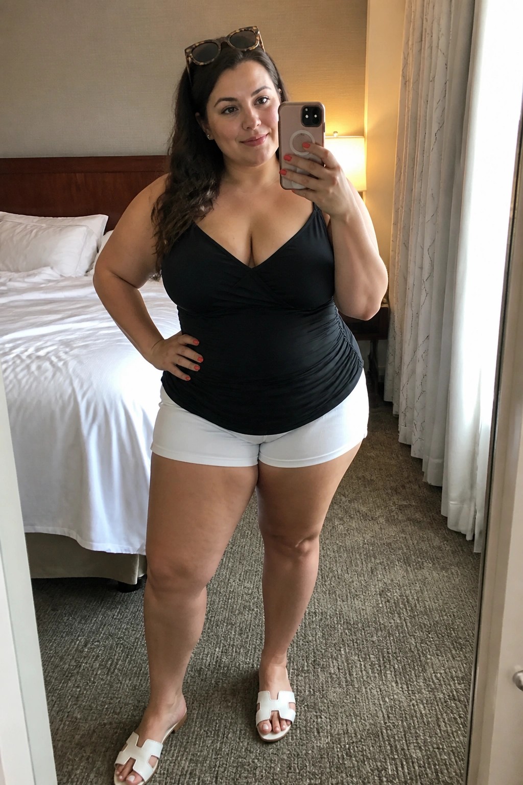 Woman in black top and white shorts taking mirror selfie in hotel room