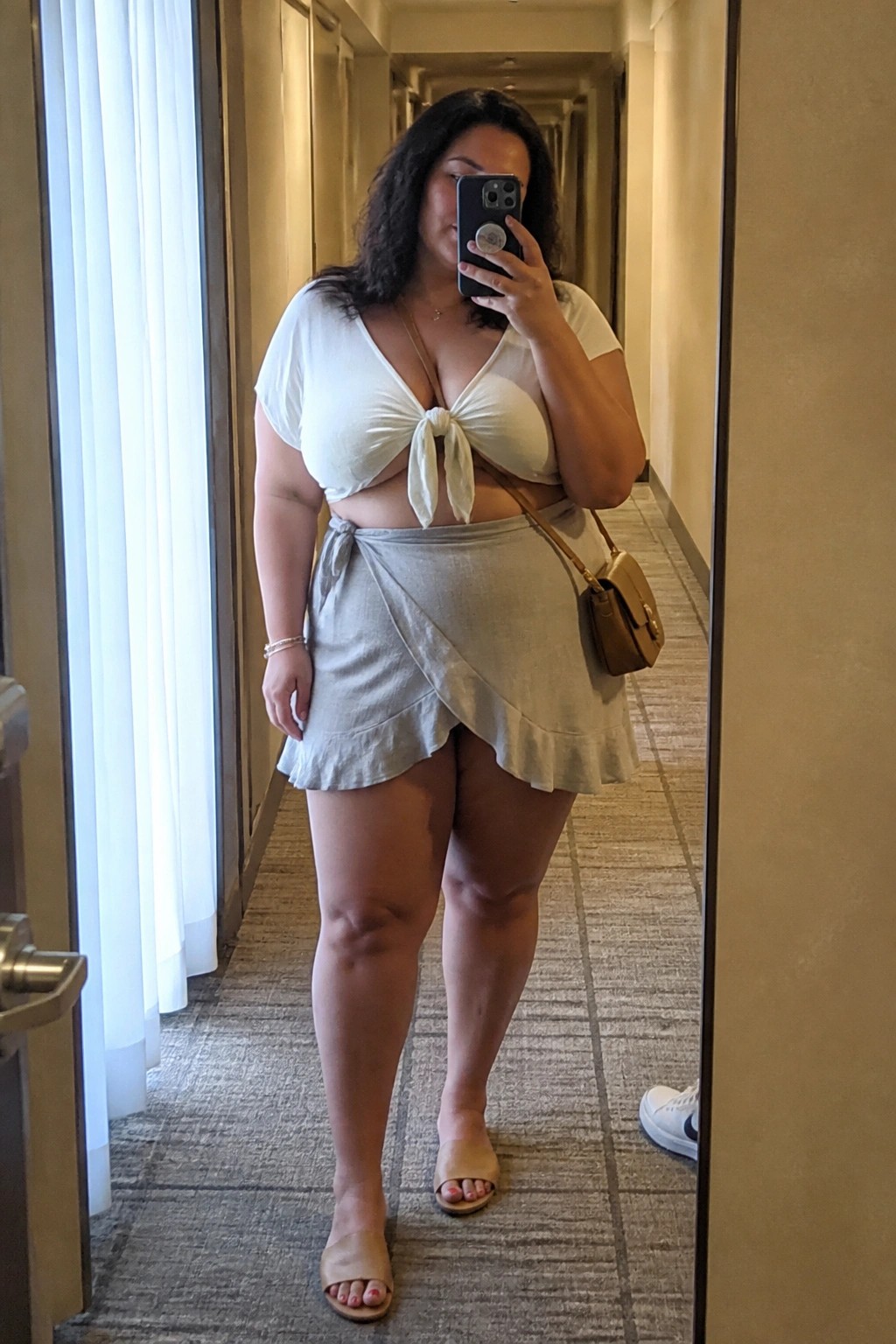 Curvy woman in white knotted crop top and gray wrap skirt taking mirror selfie in hallway