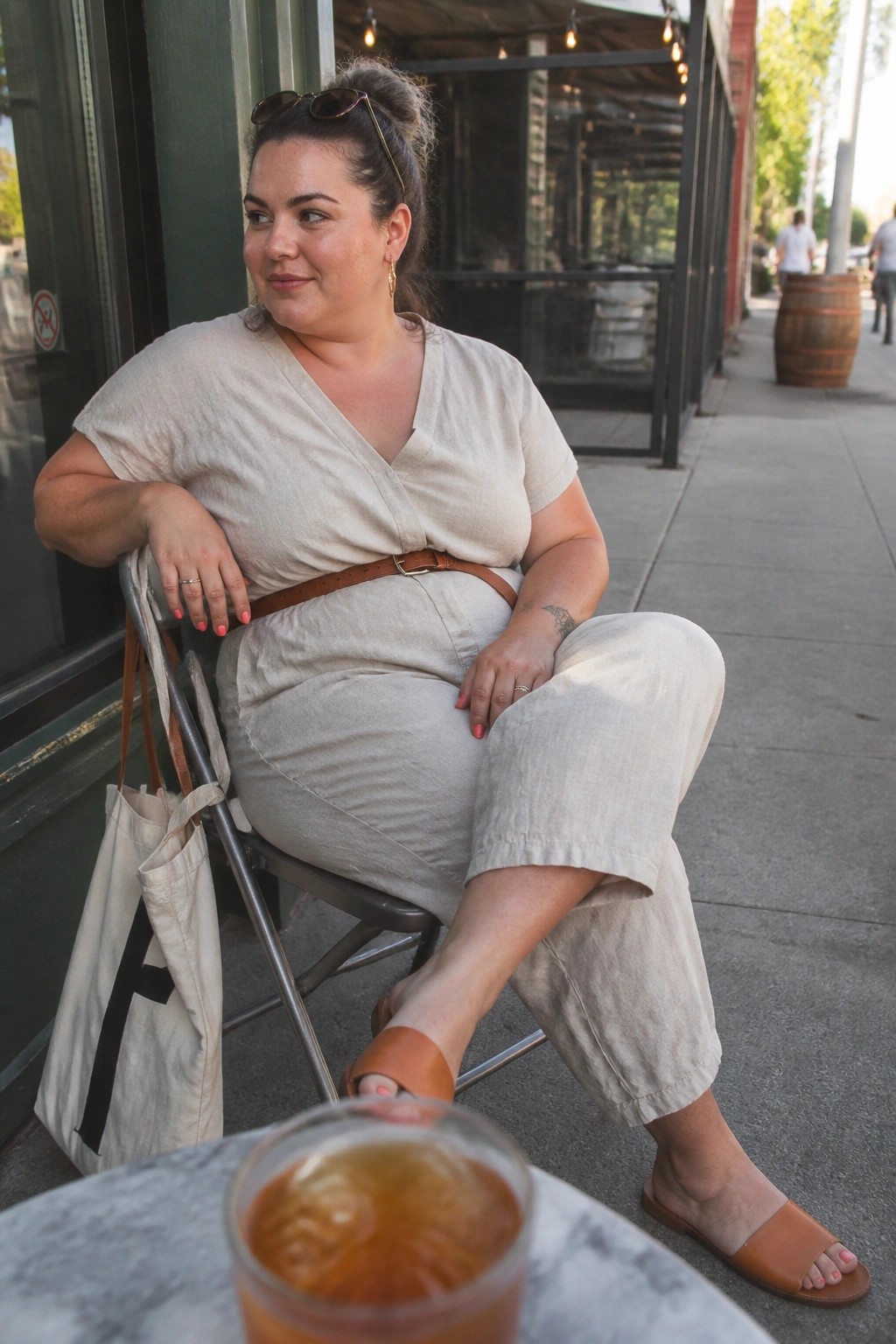 Woman in beige wrap top and pants seated outdoors by iced drink.