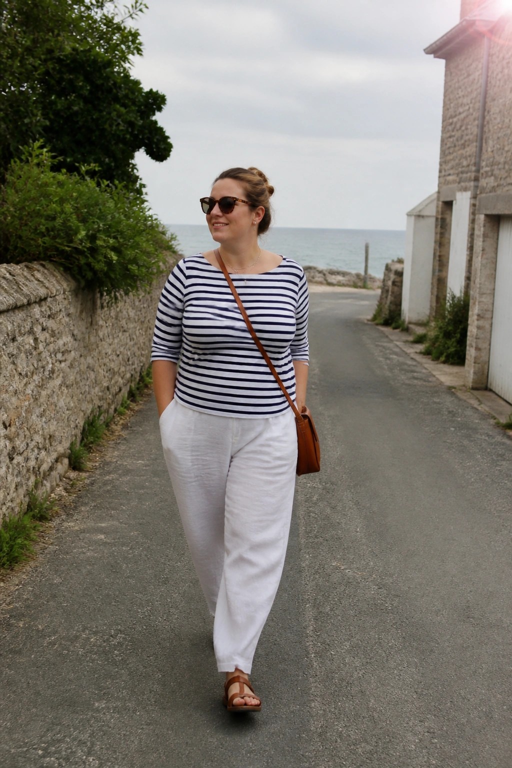 Woman in striped shirt and white pants walking on seaside path with bag