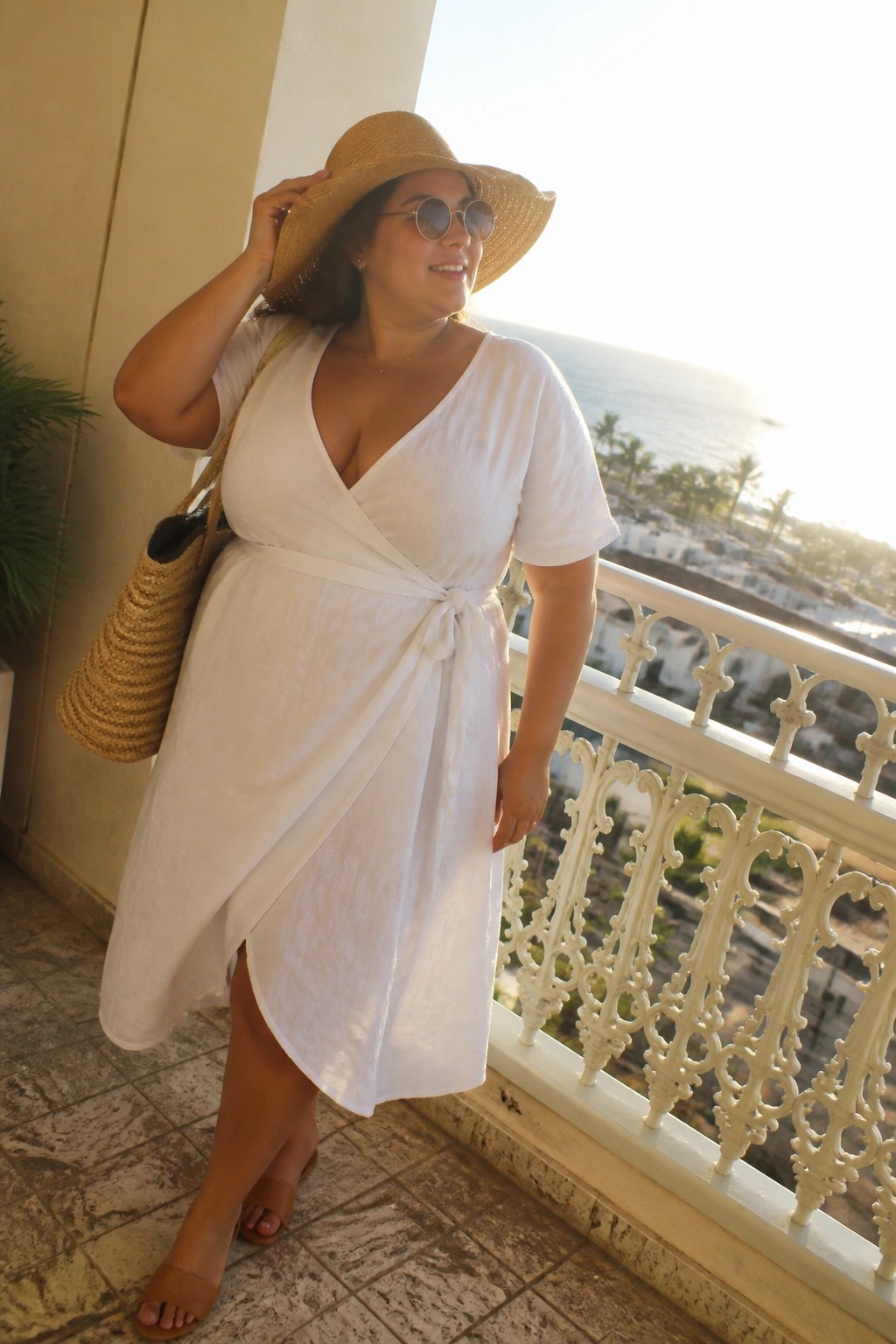 Woman in white wrap dress and straw hat on sunny ocean balcony