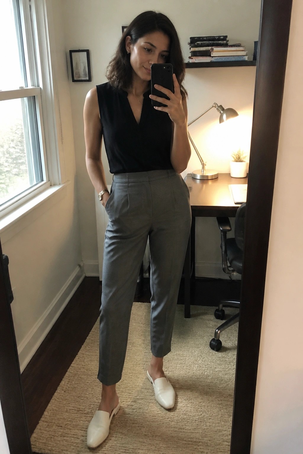 Woman in black top and gray pants taking mirror selfie in office