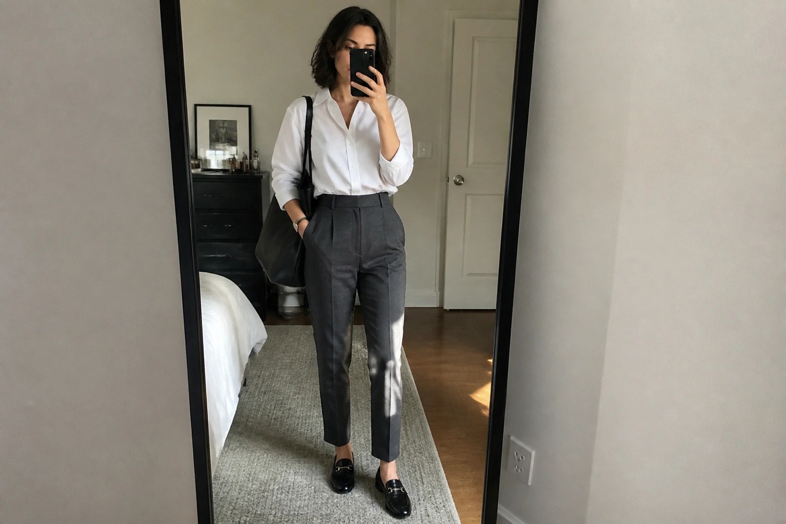 Woman in white shirt and gray trousers taking mirror selfie in bedroom