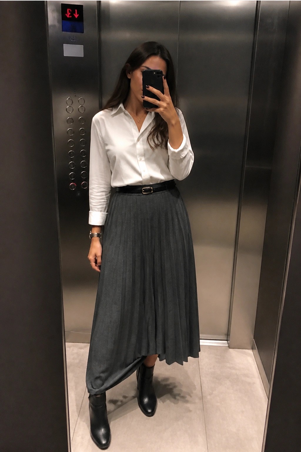 Woman in white shirt and gray pleated skirt taking selfie in elevator