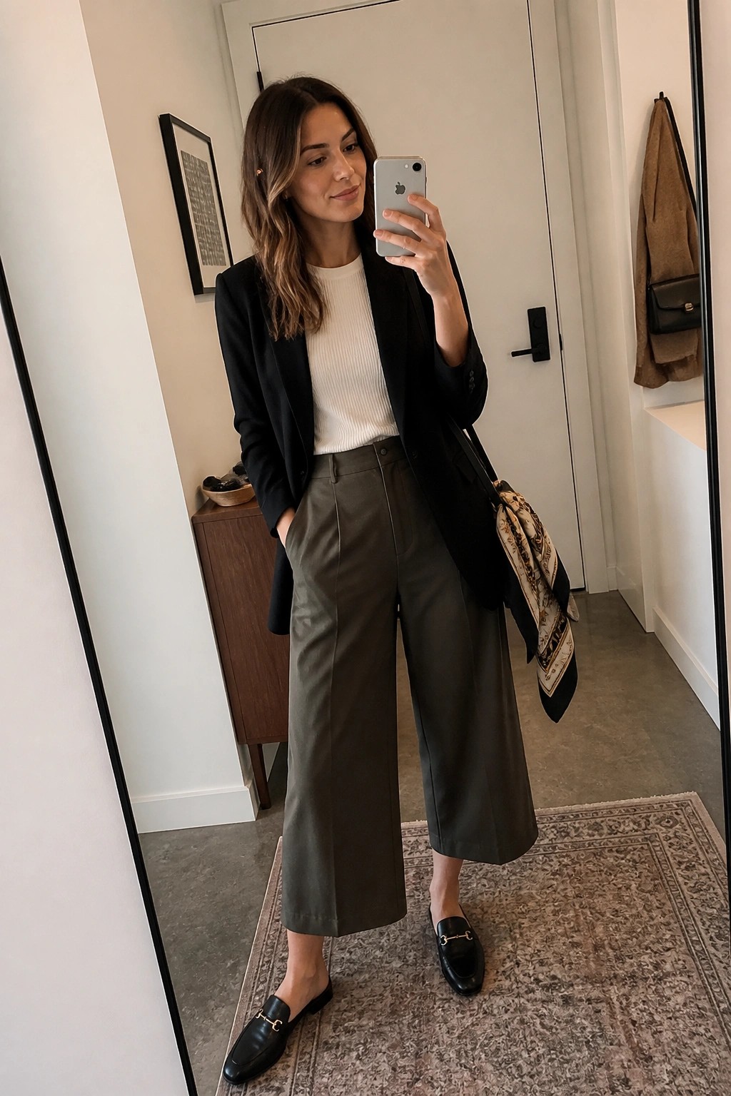 Woman in black blazer and olive trousers taking mirror selfie with iPhone