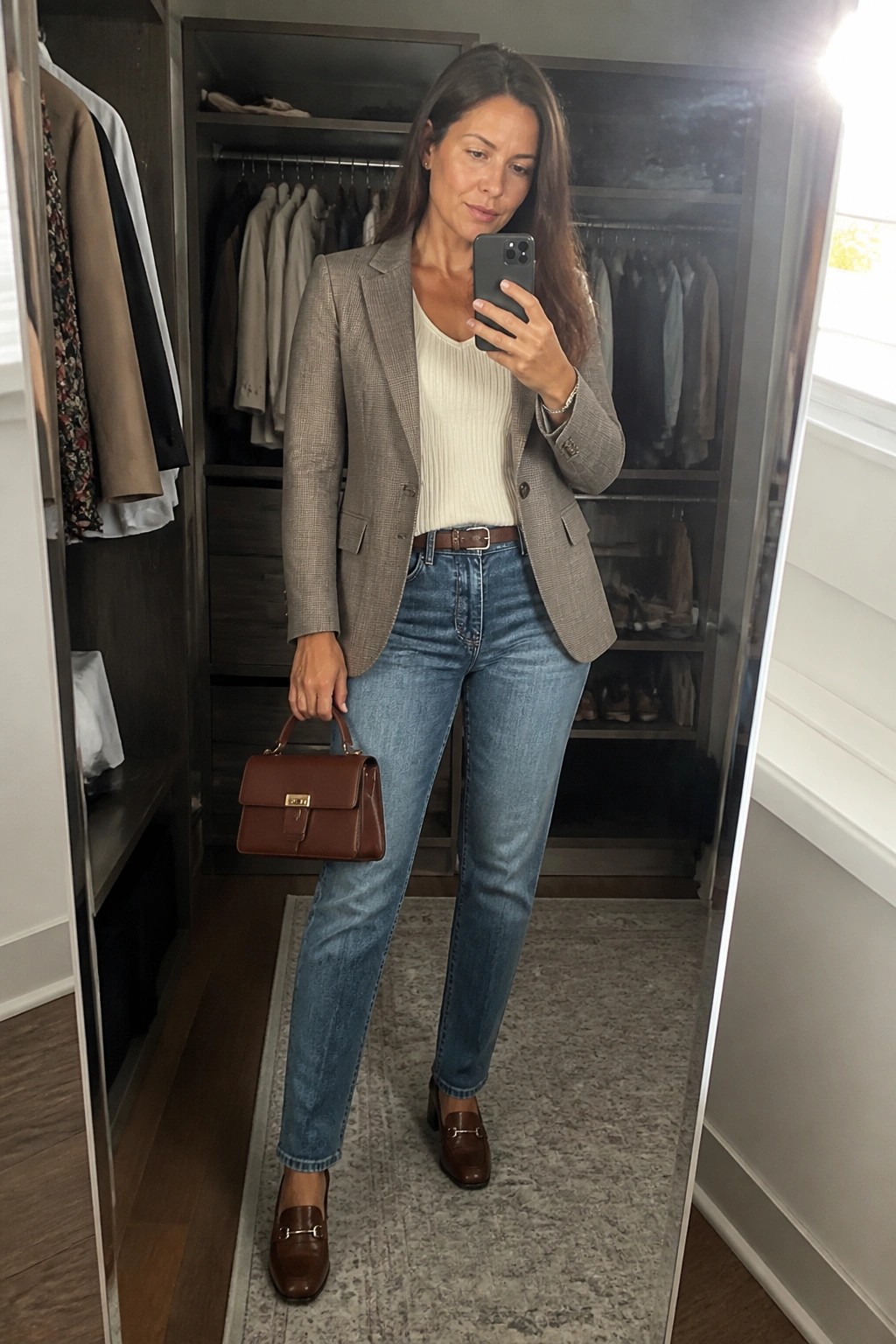 Woman wearing gray plaid blazer over cream ribbed sweater with blue jeans brown leather belt and matching loafers plus handbag while holding phone for mirror selfie in closet