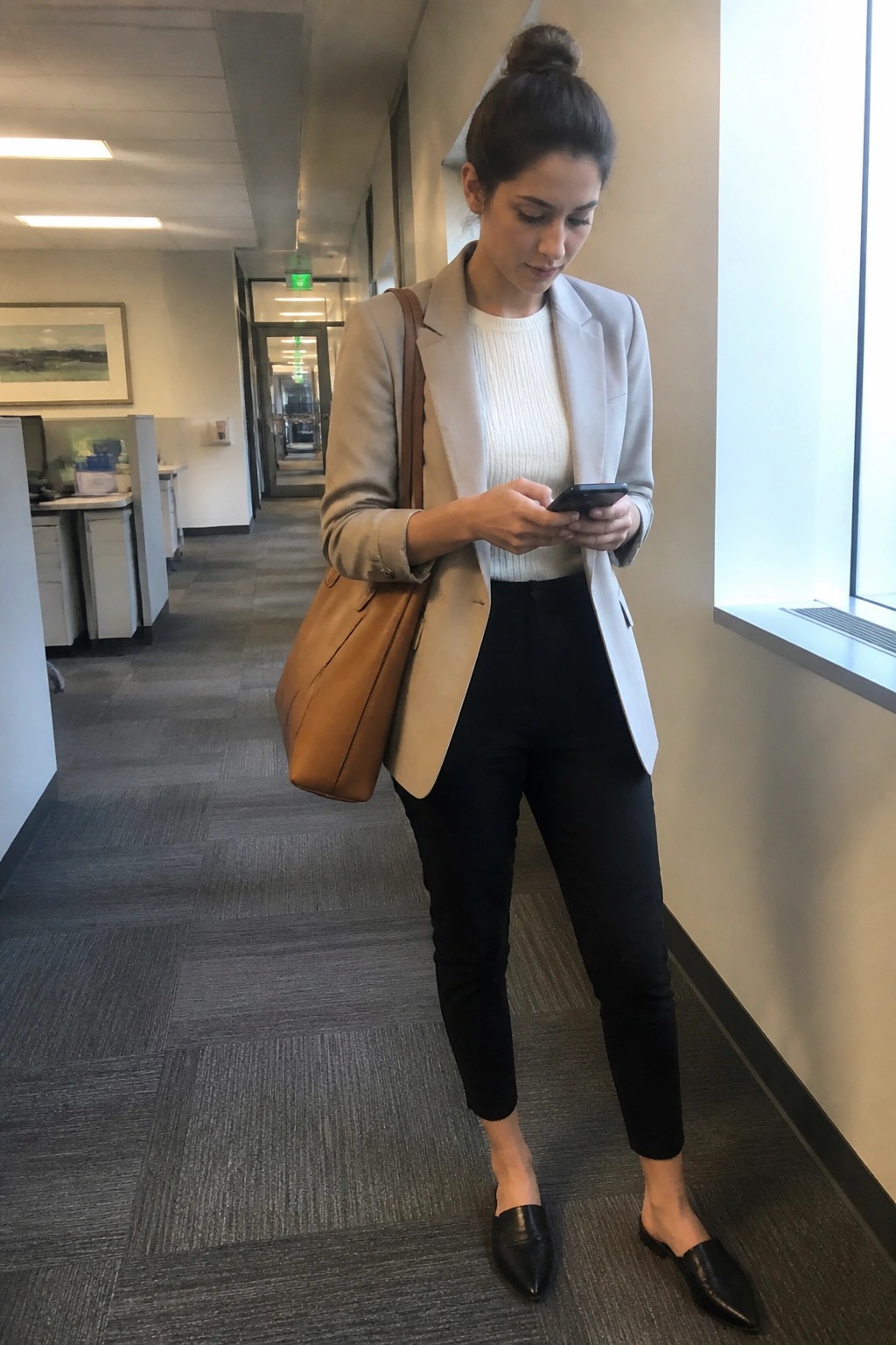 Woman standing in office hallway wearing a beige blazer over white ribbed sweater black slim pants and black pointed mules while holding a phone with brown leather tote on shoulder