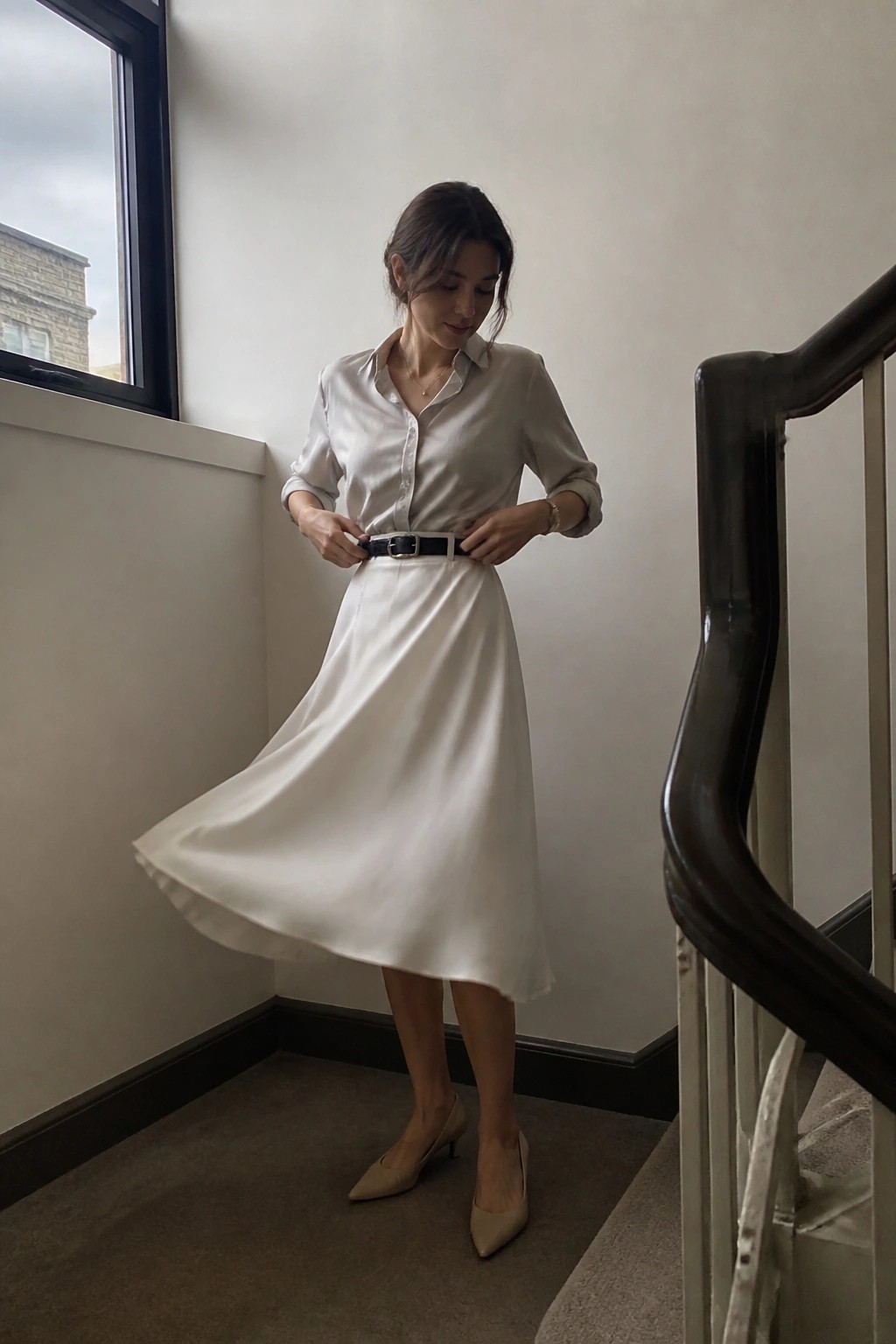 Woman wearing a light beige satin button-up shirt tucked into a matching white satin midi skirt, cinched with a black belt, paired with nude pointed kitten heels and delicate gold jewelry.