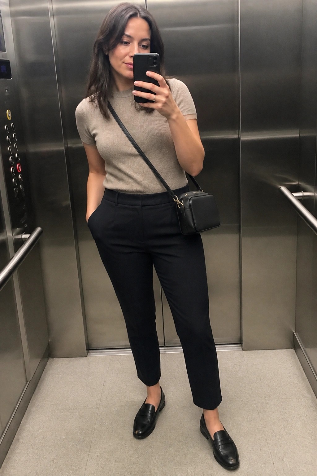 Woman in beige top and black pants taking mirror selfie in elevator