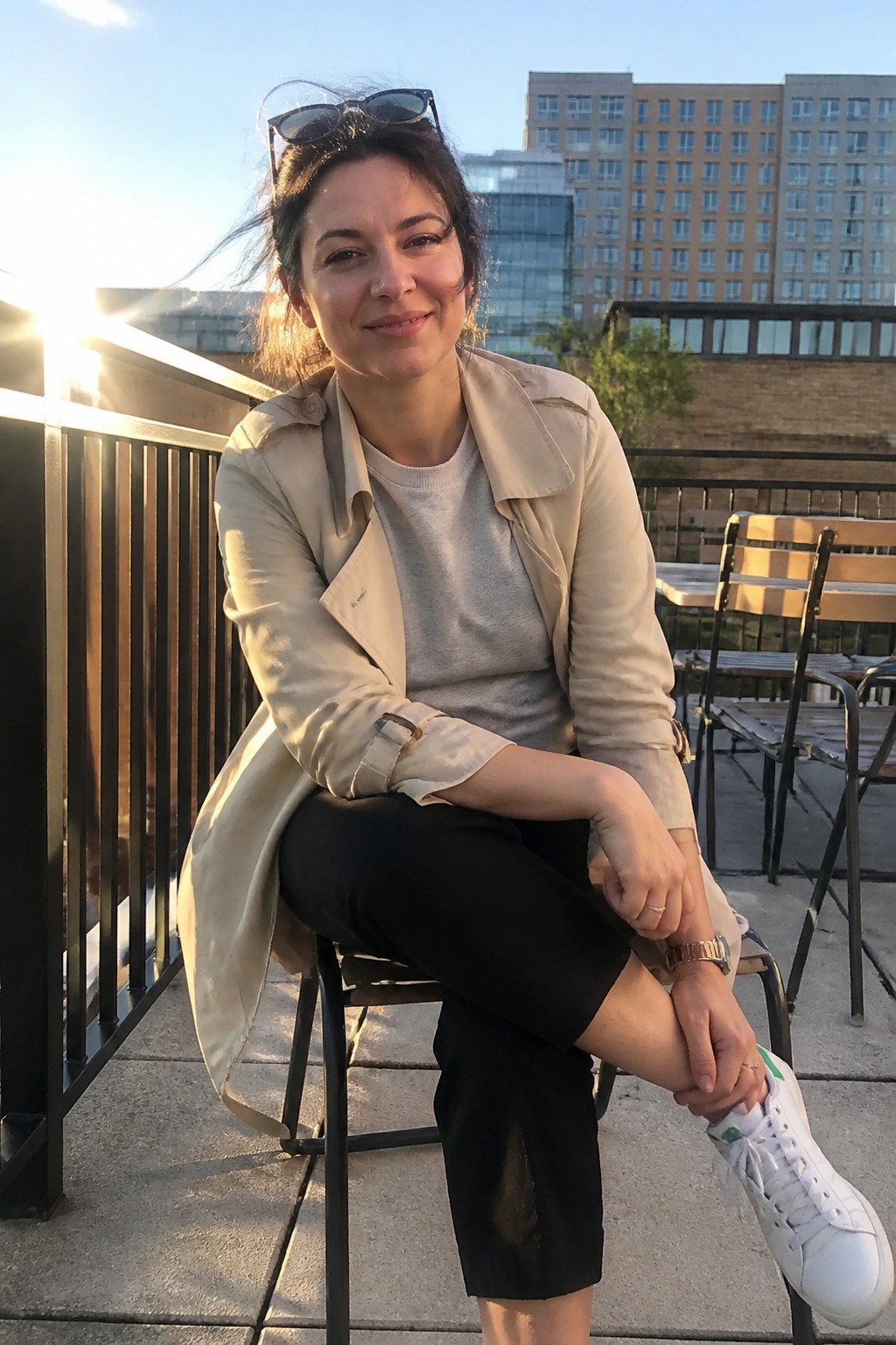 Smiling woman in beige trench coat with sunglasses on head sits on city rooftop