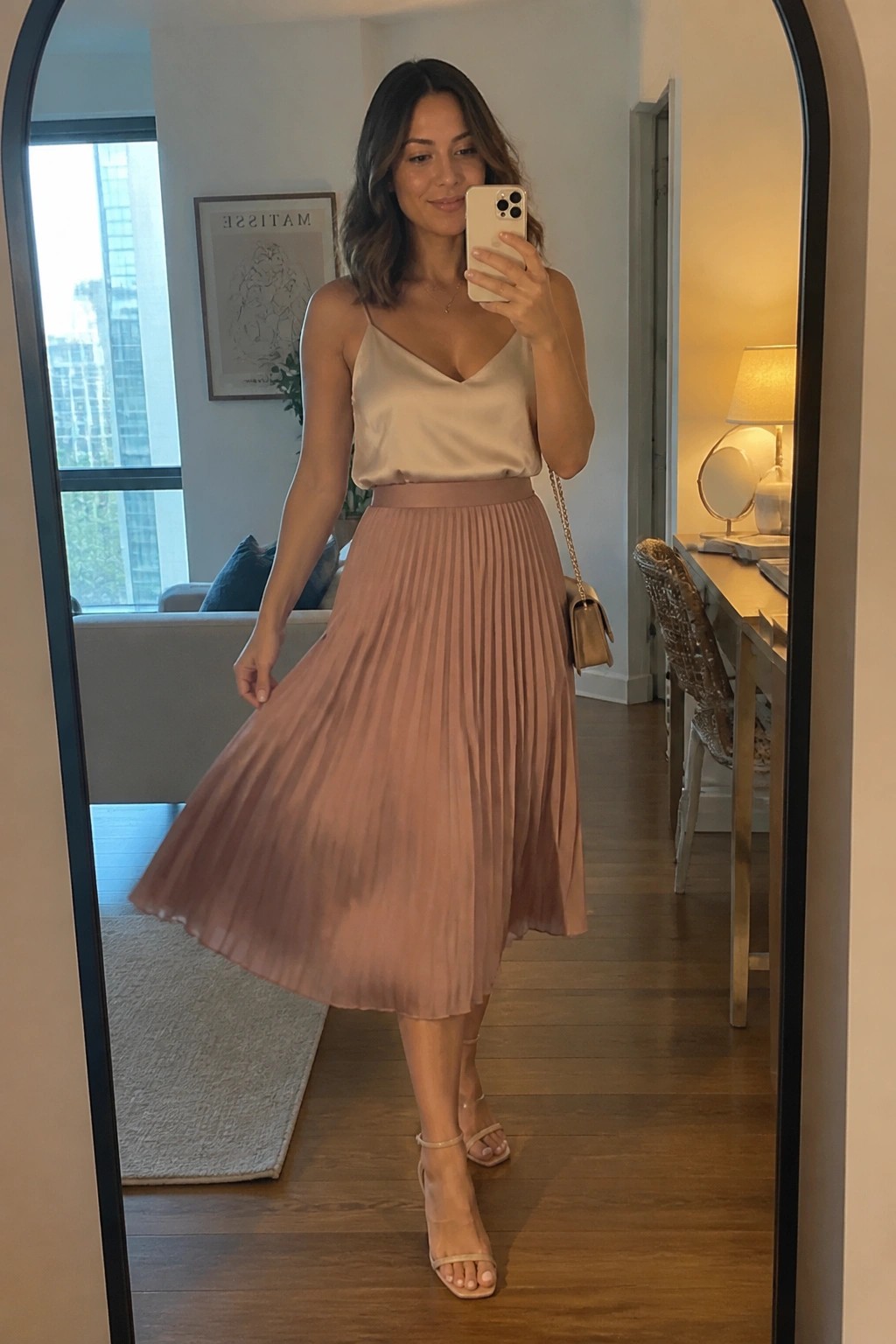 Woman wearing a beige satin camisole top with thin straps and a blush pink pleated midi skirt paired with nude strappy heels and a small beige purse while taking a mirror selfie.
