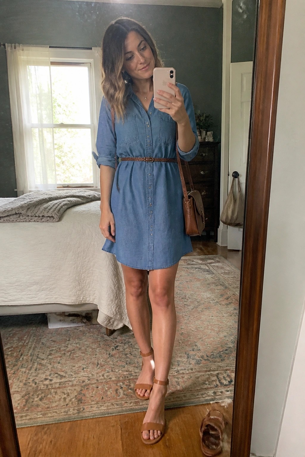 Woman in blue denim shirt dress taking mirror selfie in bedroom with brown belt and sandals