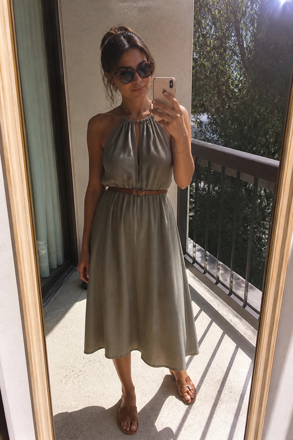 Woman in sage green halter dress with brown belt taking balcony selfie.