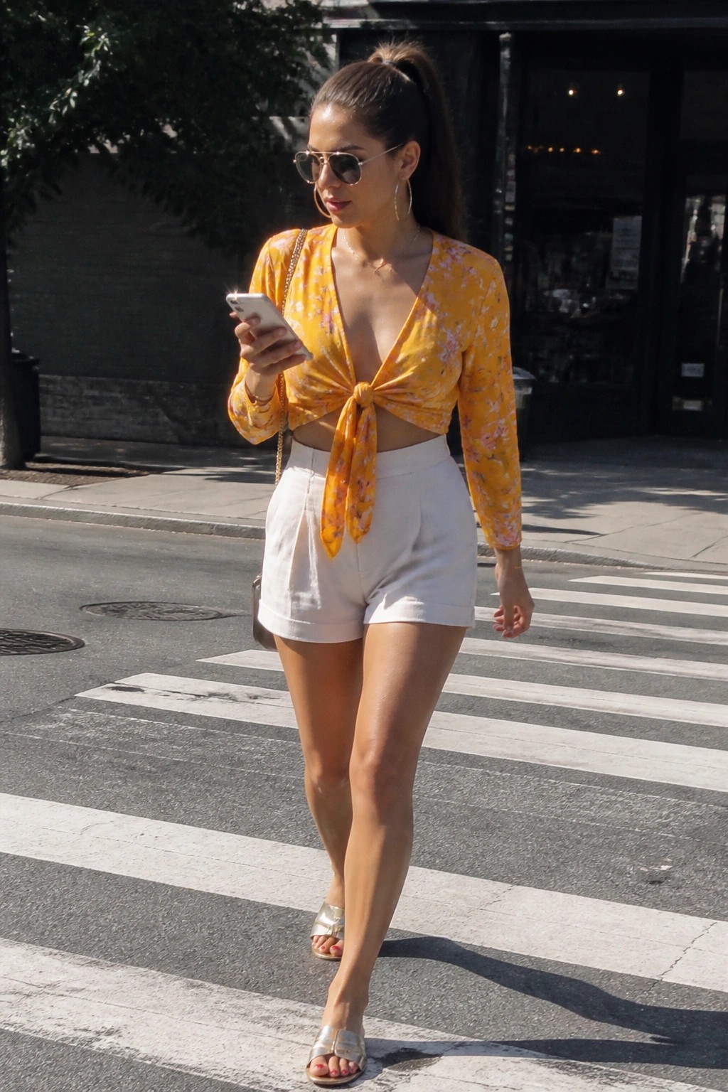Woman in yellow floral knotted top and white shorts checking phone on crosswalk