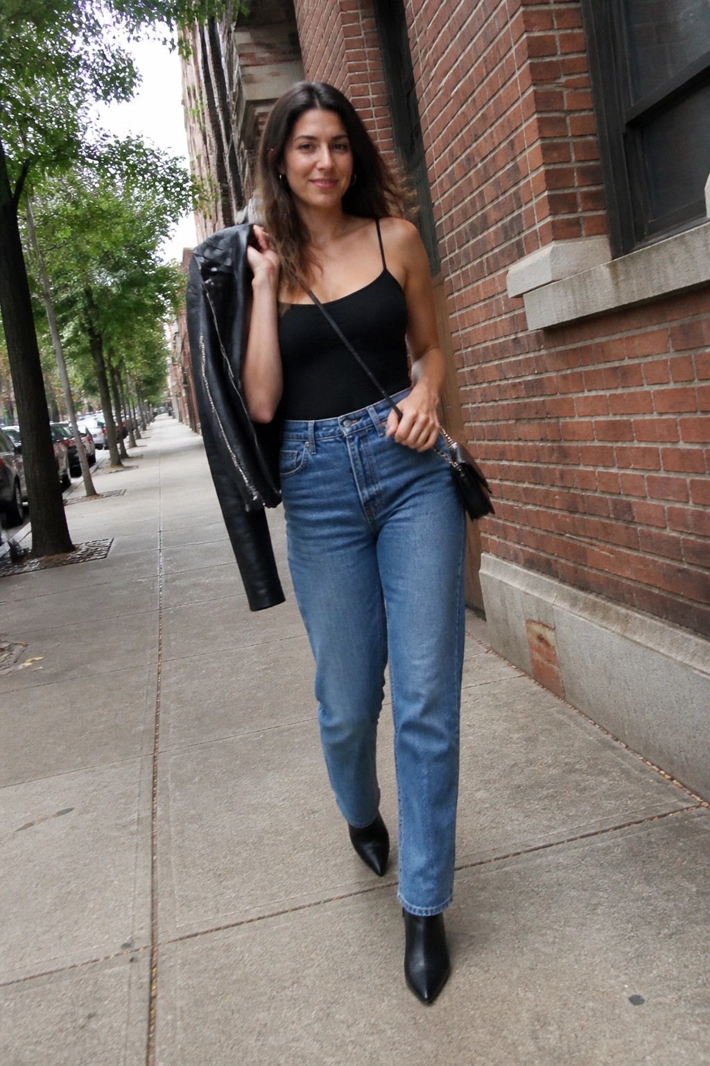 Woman wearing a black camisole tucked into blue straight-leg jeans, black pointed ankle boots, with a black leather jacket draped over one shoulder and a small black crossbody bag.