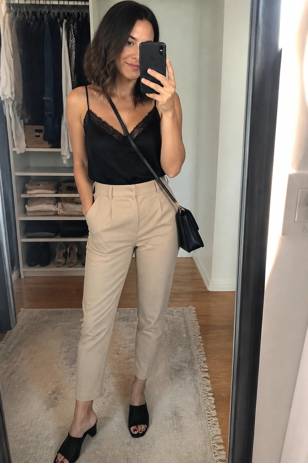 Woman in black satin camisole with lace trim, beige high-waisted trousers, black crossbody bag, and black heeled mules posing for a mirror selfie in a closet area.
