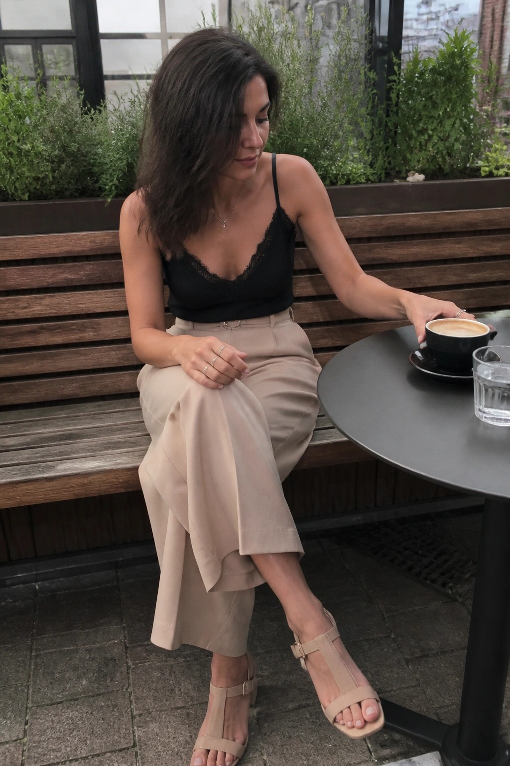 Woman in black lace top and beige pants seated at outdoor cafe with coffee.
