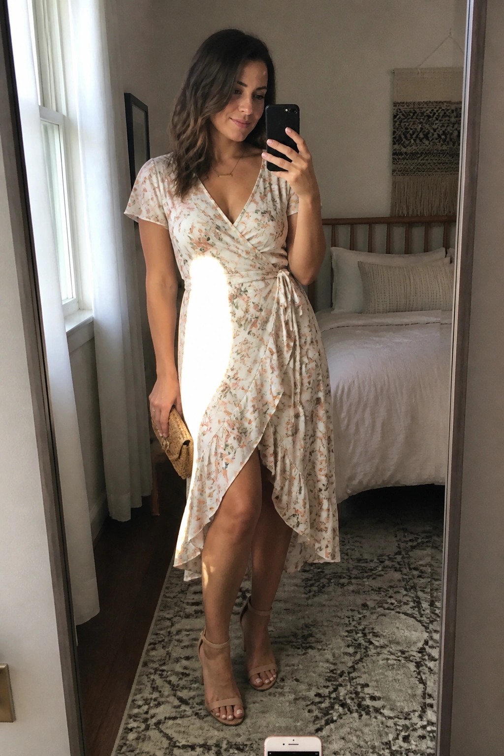 Woman wearing a short sleeve floral wrap dress in light cream with peach accents, side tie, asymmetrical ruffled hem, nude strappy heels, and holding a woven clutch while taking a mirror selfie.