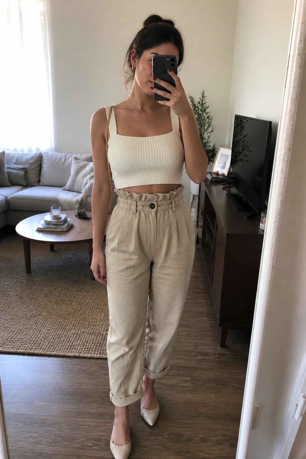Woman in white crop top and beige paperbag pants taking mirror selfie in living room