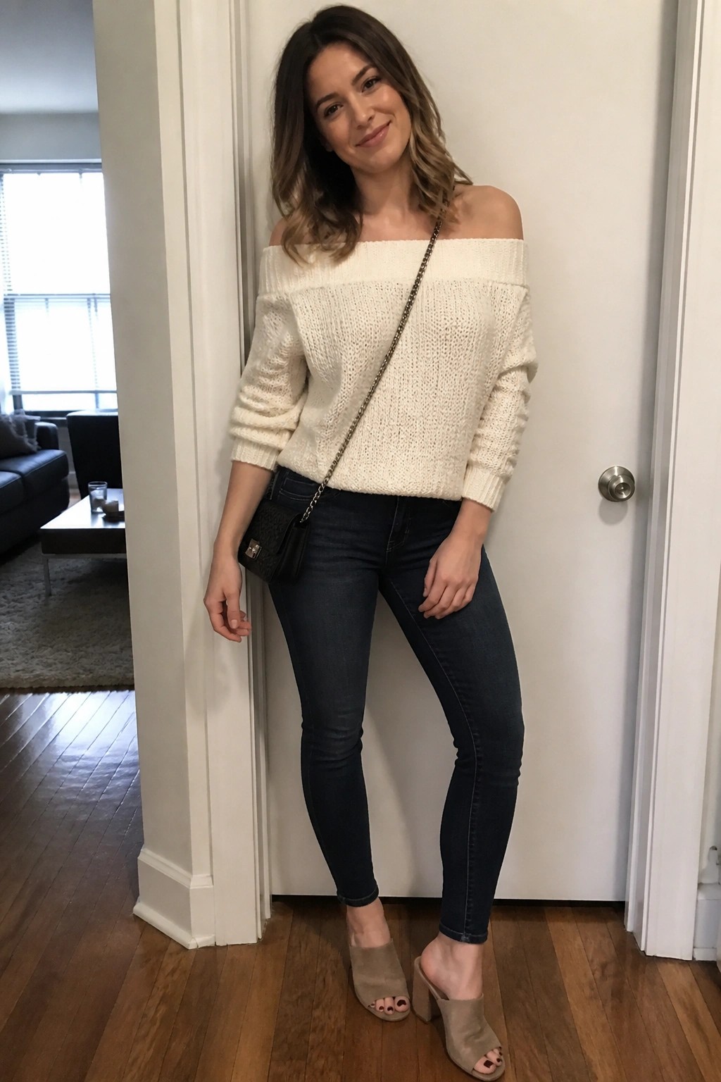 Woman in off-shoulder cream knit sweater, dark skinny jeans, beige open-toe block heel mules, and black chain strap crossbody bag standing indoors.