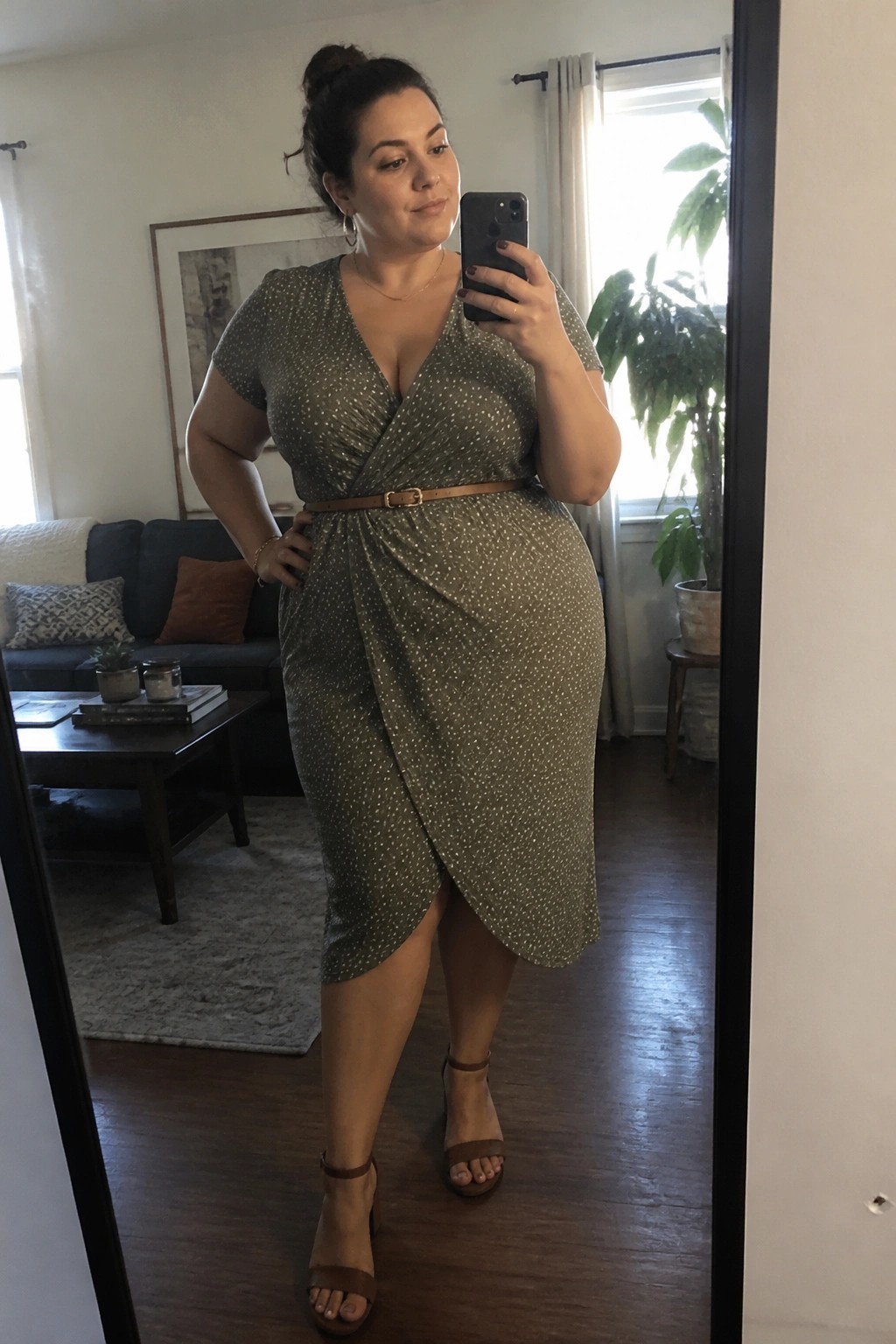 Woman in green polka-dot wrap dress taking mirror selfie in living room