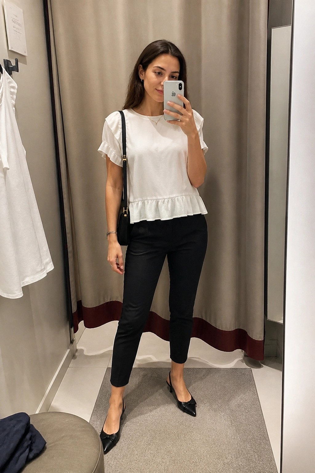 Woman in white ruffled peplum top and black pants taking mirror selfie in fitting room