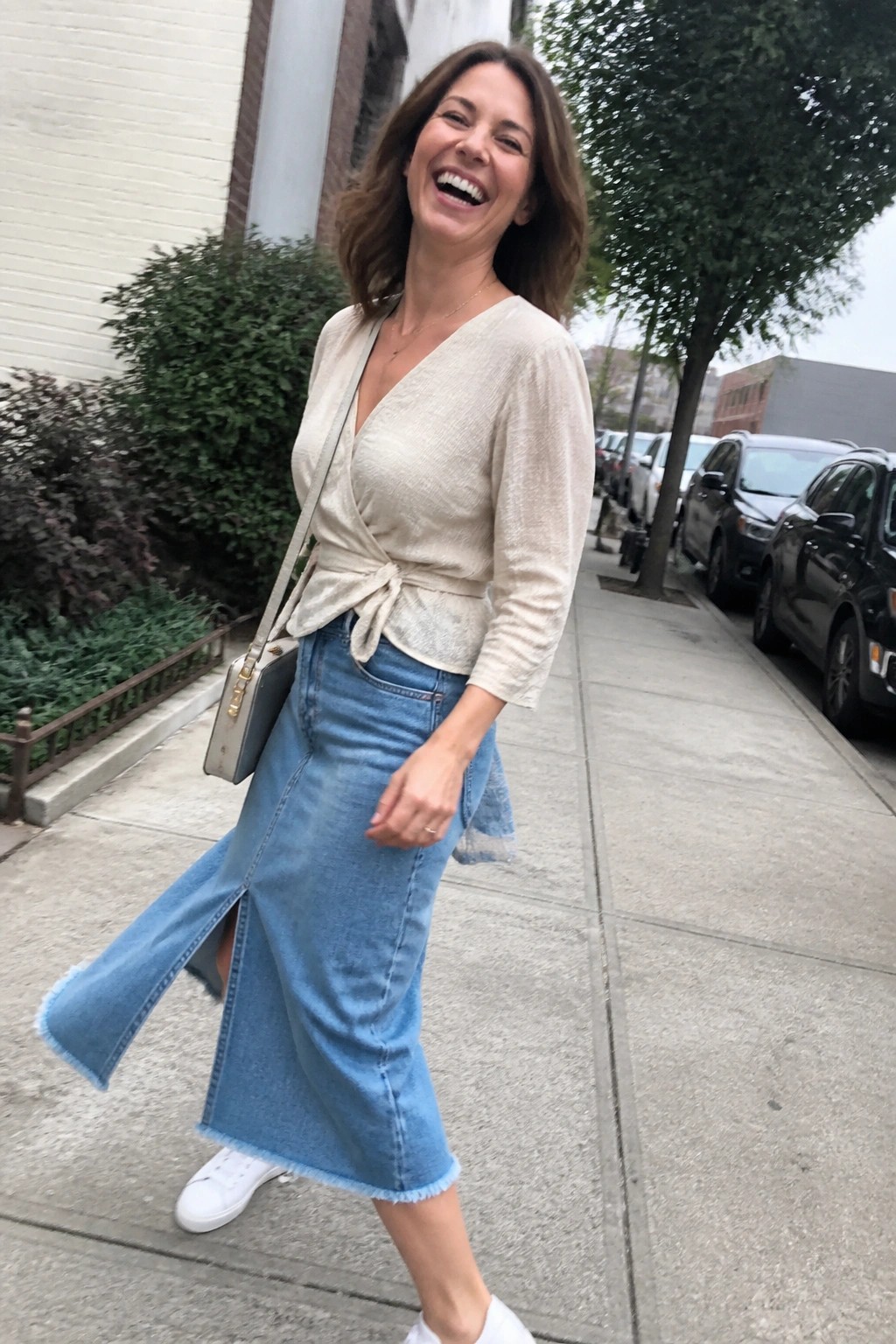 Woman in beige wrap top and denim midi skirt laughs on sidewalk