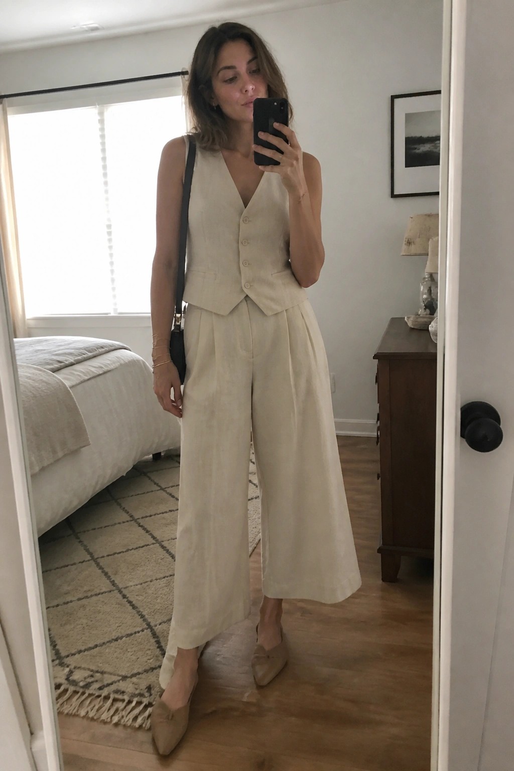 Woman in beige linen vest and wide-leg pants taking bedroom mirror selfie