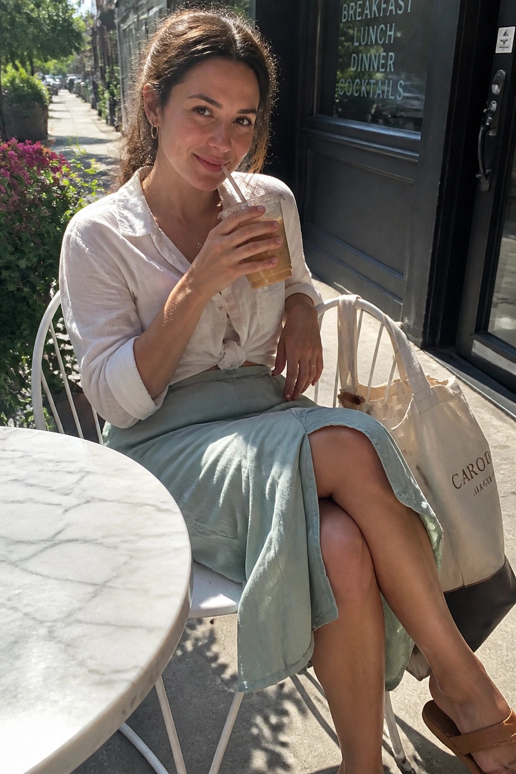 Woman seated outdoors wearing a tied white button down shirt and sage green midi skirt with brown sandals and a canvas tote bag while holding an iced coffee.