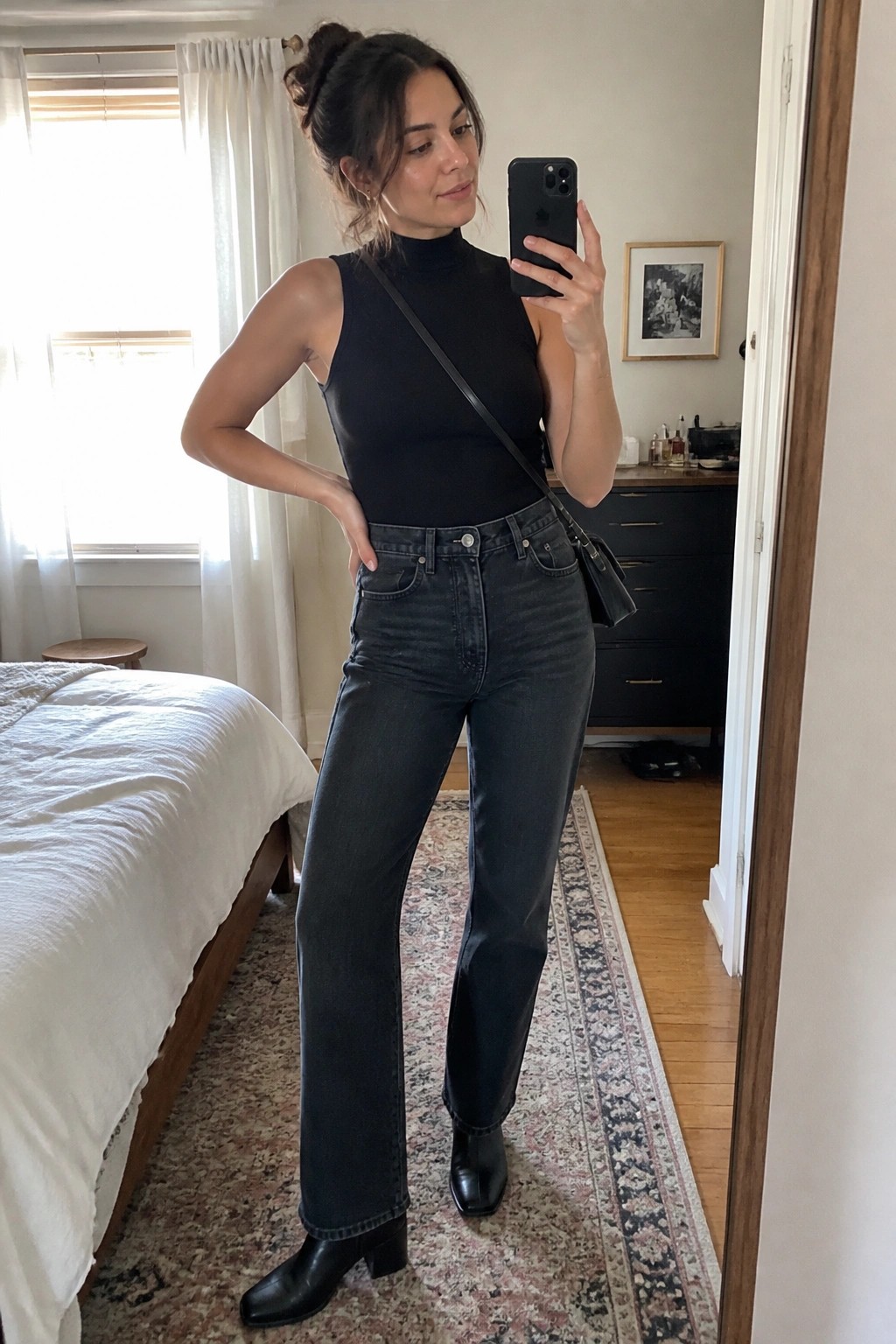 Woman wearing a black sleeveless turtleneck bodysuit with dark straight leg jeans and black heeled ankle boots while holding a phone for a mirror selfie