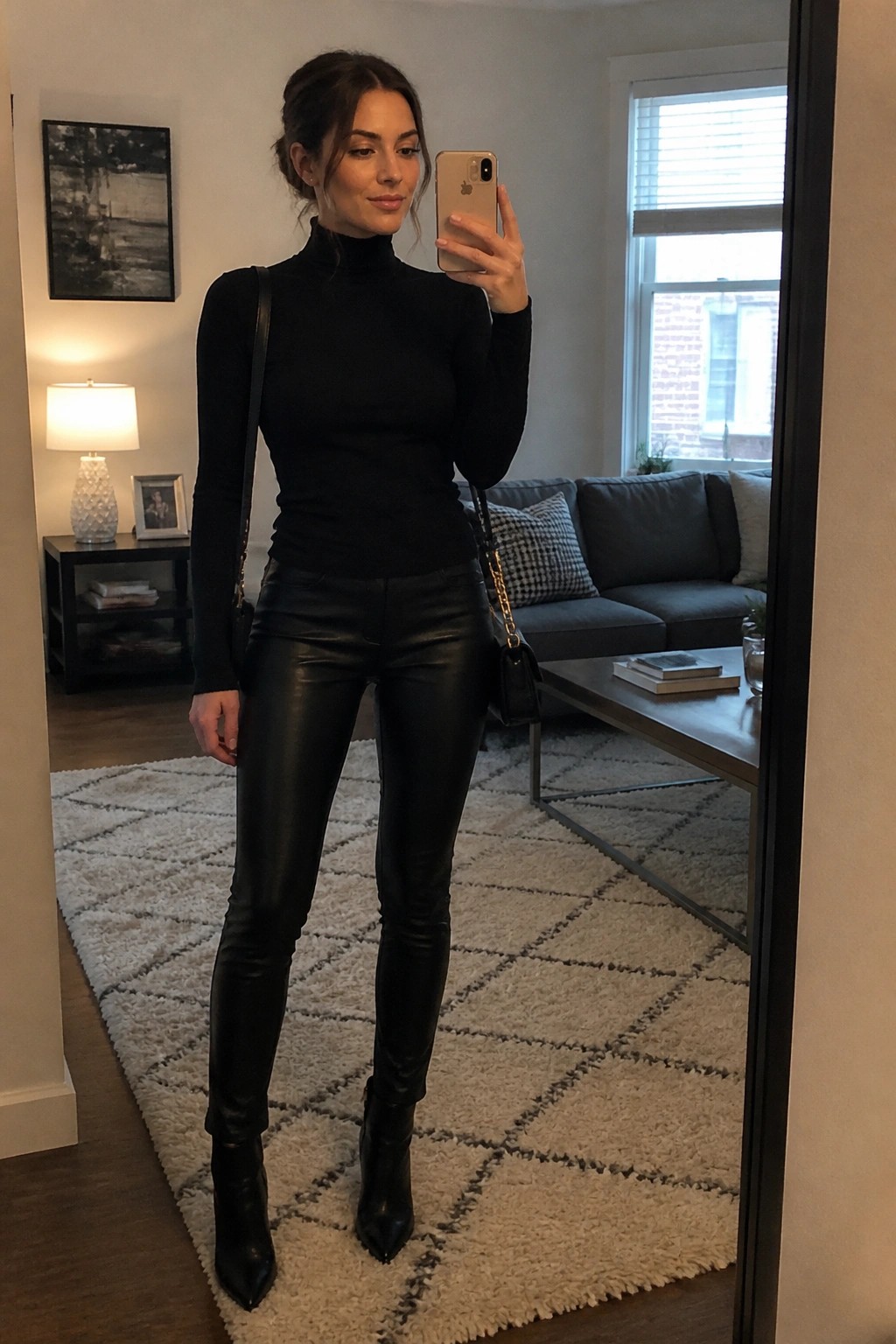 Woman wearing a black turtleneck top, black leather pants, and black pointed ankle boots while holding a phone for a mirror selfie in a living room setting.
