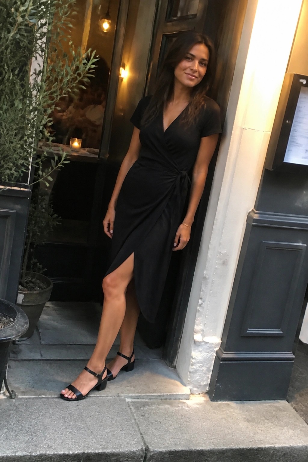 Woman wearing a black short sleeve wrap midi dress tied at the waist with a front slit paired with black block heel sandals standing near a doorway.