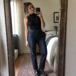 Woman wearing a black sleeveless turtleneck bodysuit with dark straight leg jeans and black heeled ankle boots while holding a phone for a mirror selfie