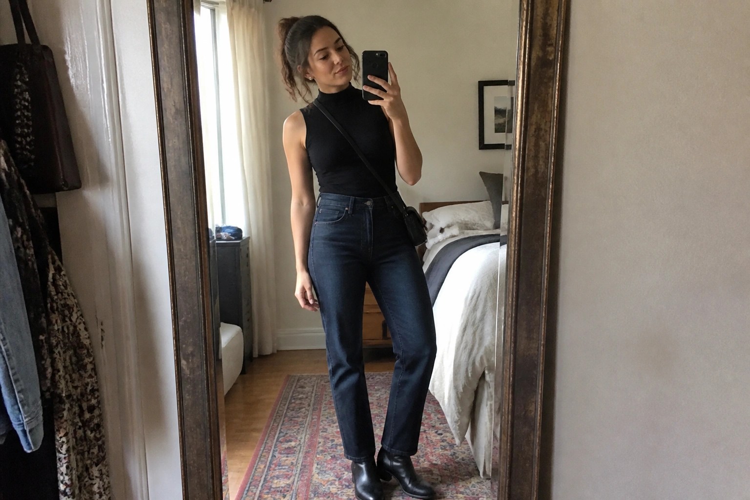 Woman wearing a black sleeveless turtleneck bodysuit with dark straight leg jeans and black heeled ankle boots while holding a phone for a mirror selfie