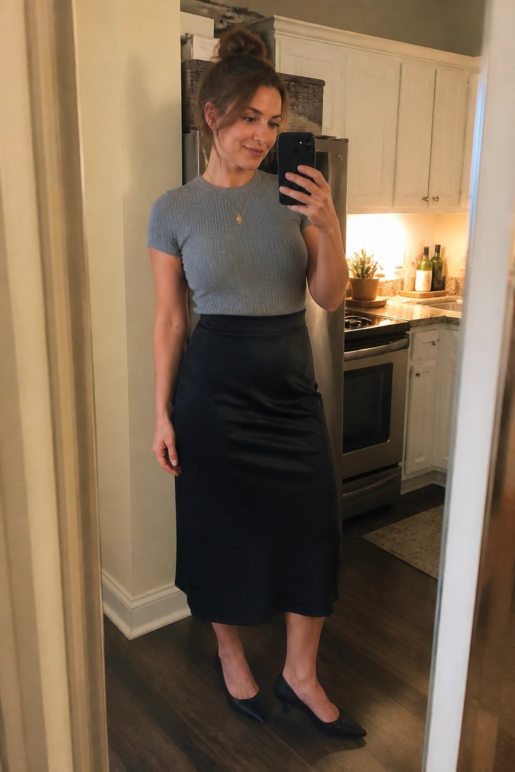 Woman in gray top and black skirt taking mirror selfie in kitchen